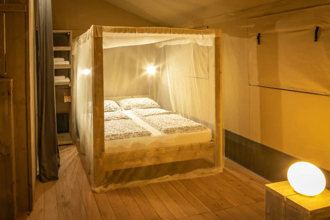 Camera da letto del Wood Lodge per 2-6 persone con letto a baldacchino sotto una zanzariera, armadio a vista con asciugamani e una lampada sul comodino