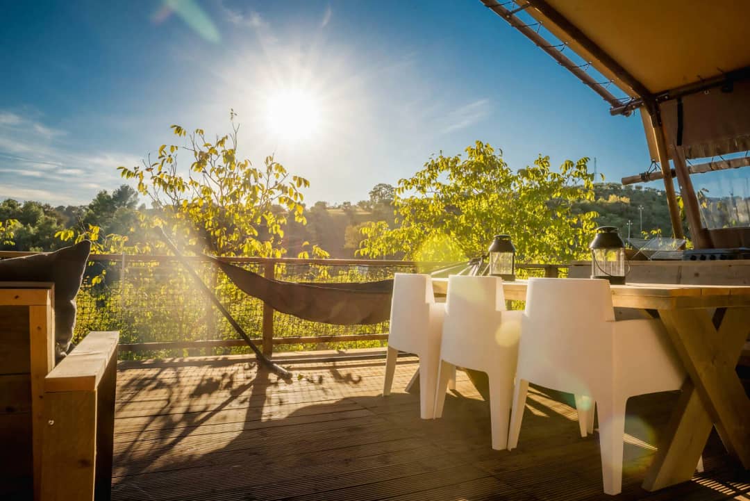 Tavolo da pranzo in legno con gambe a croce e sedie bianche sulla terrazza del Desert Lodge XL, con il sole che splende sopra gli alberi
