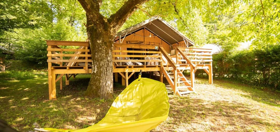 Safari Lodge su palafitte sotto un grande albero, con una scala di legno che porta alla terrazza e un'amaca gialla in primo piano