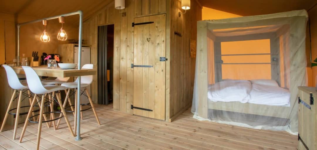 Zona giorno del Desert Lodge XL con bancone in legno, sgabelli da bar bianchi, porta in legno e letto a baldacchino con zanzariera
