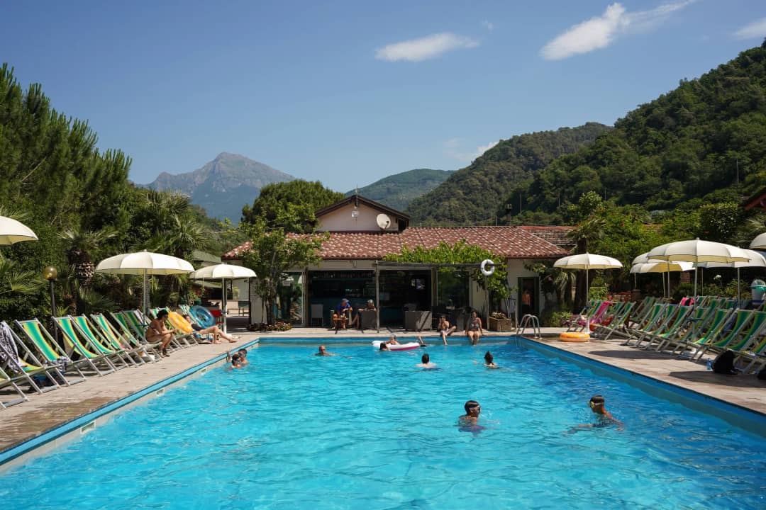 La piscina del Camping Delle Rose con sedie a sdraio verde e bianche, ombrelloni bianchi e una vetta in sottofondo