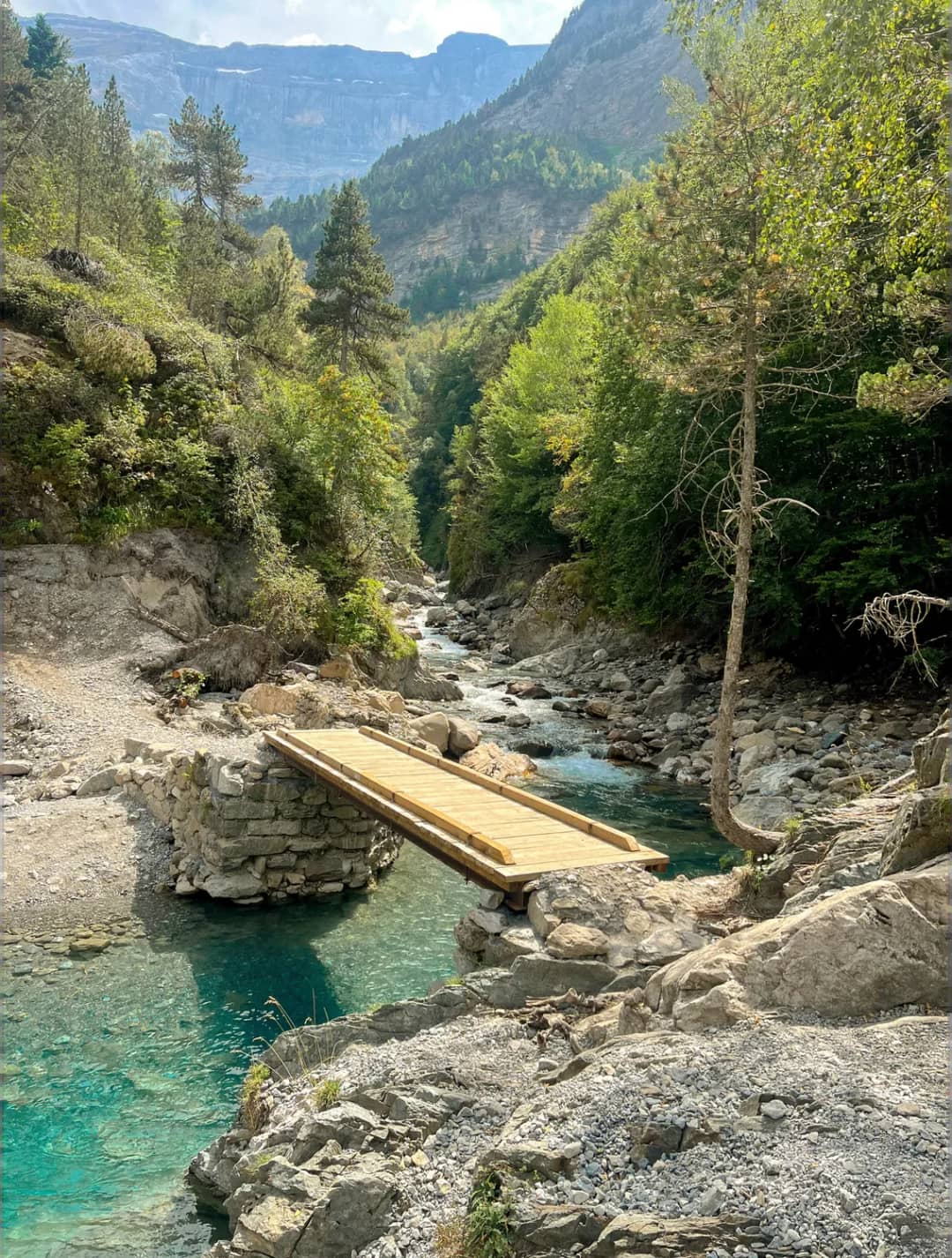Torrente di montagna azzurro e limpido con piattaforma in legno in una valle boscosa circondata da rocce e conifere, con montagne sullo sfondo.